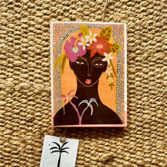Decorative tile with a stylized illustration of a woman with flowers on her head, placed on a textured surface.