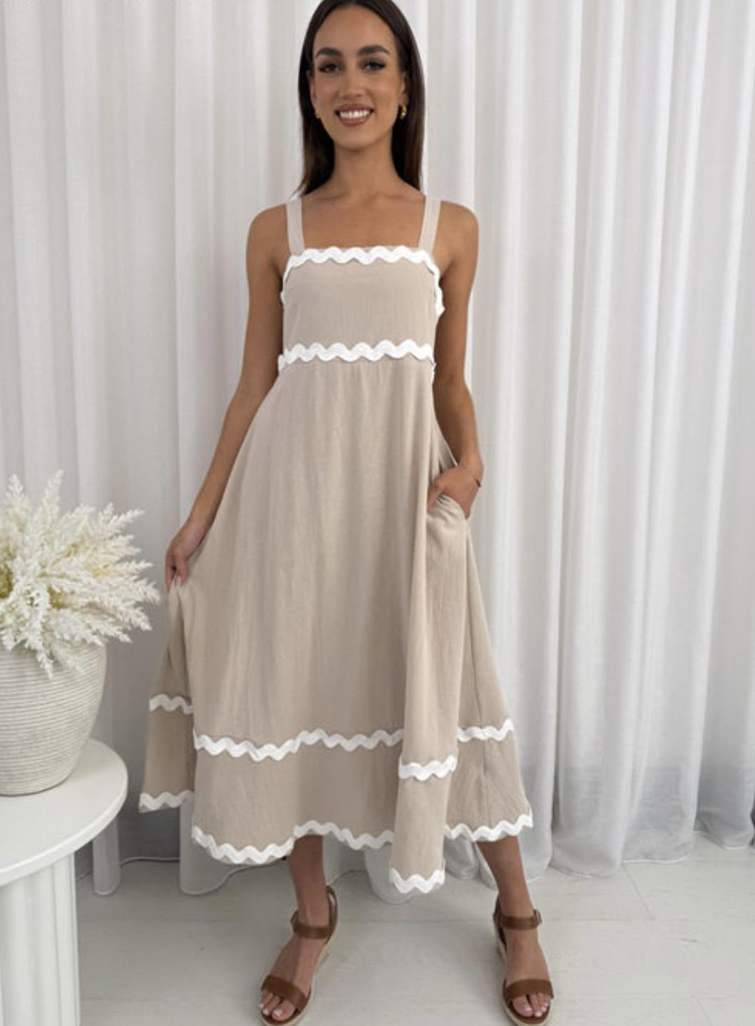 Woman wearing a beige dress with white trim against a white curtain background
