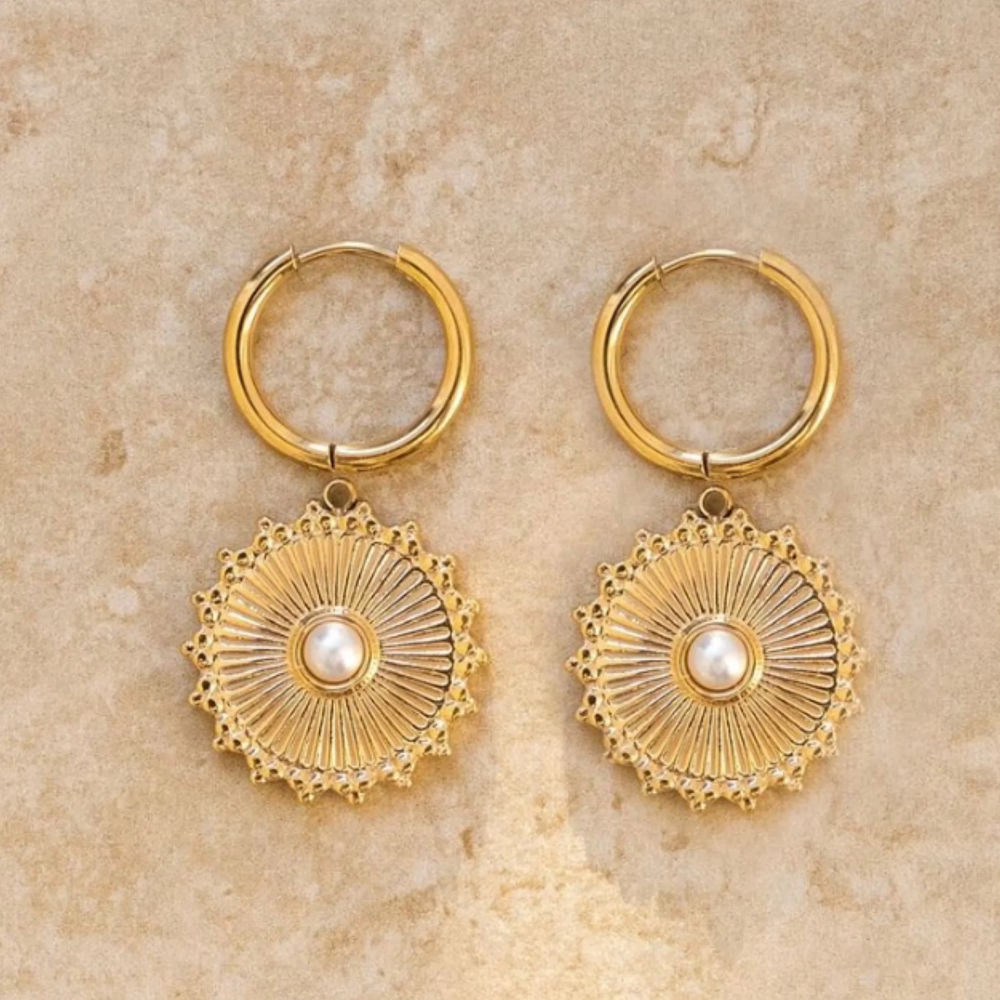 Gold hoop earrings with circular designs and pearl-like centers on a beige stone background