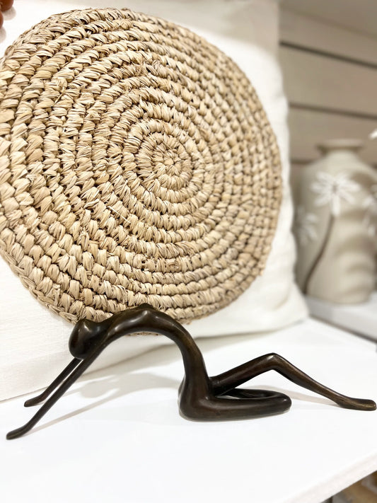 Decorative rattan round pillow with a brass lady posing sculpture on a white surface.