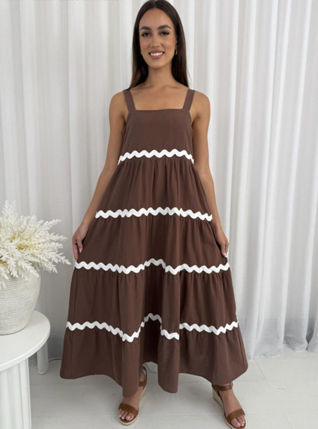 Woman wearing a brown dress with white ruffle details against a white curtain background