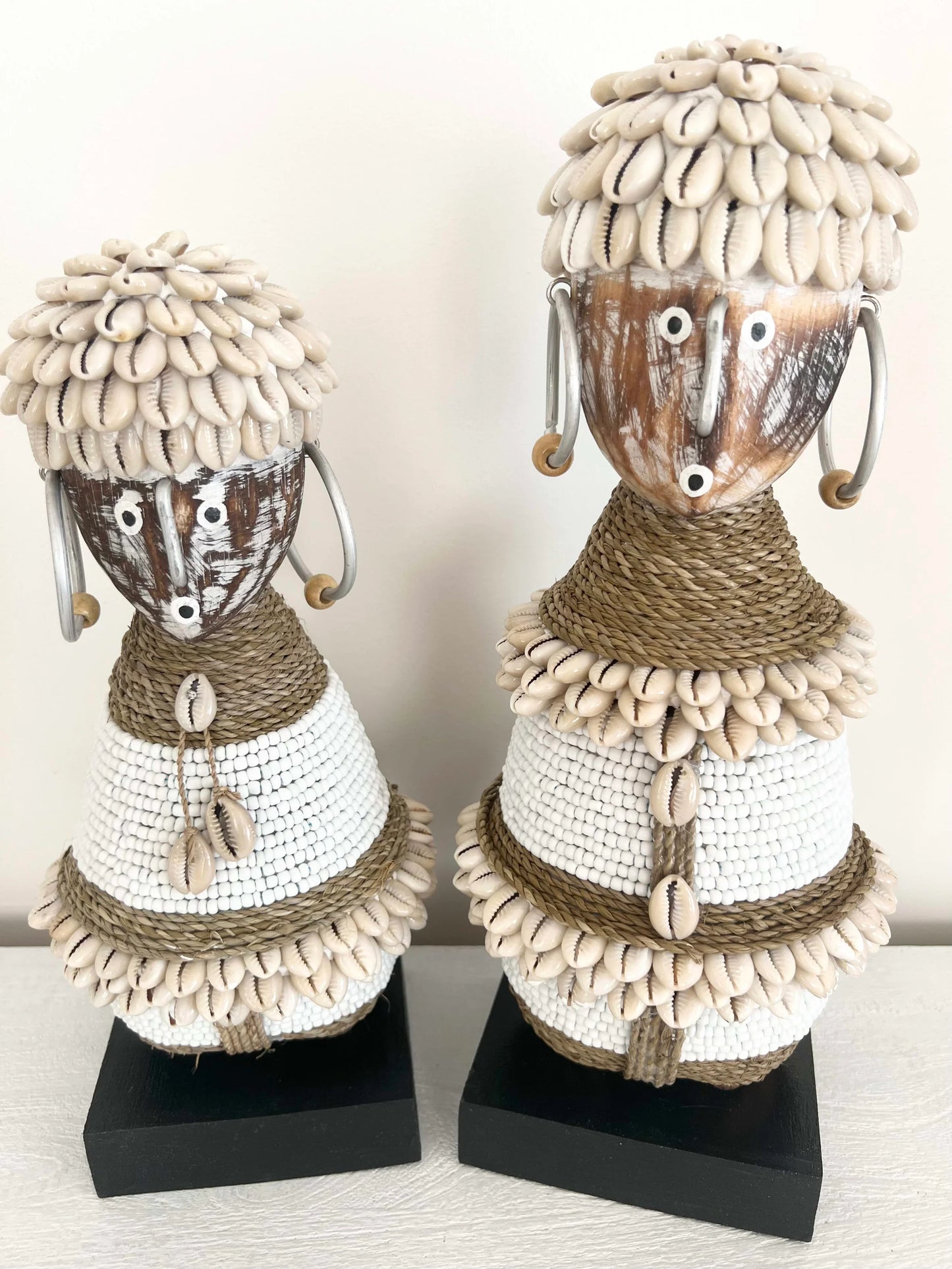 Two decorative figures made of shells and rope on black stands against a white background