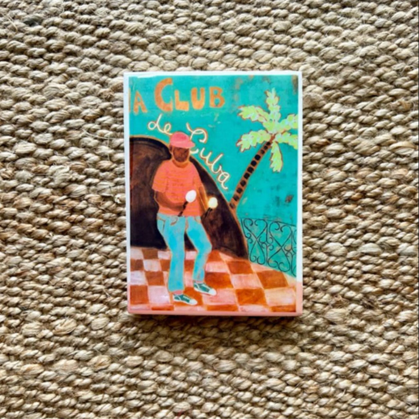 Decorative tile with a design of a man in a red hat and orange shirt on a checkered floor, palm tree, and text 'A Club de Cuba' on a textured beige background.
