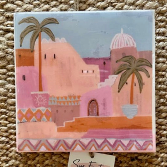 Decorative tile with a pink building and palm trees on a blue background, placed on a textured surface.
