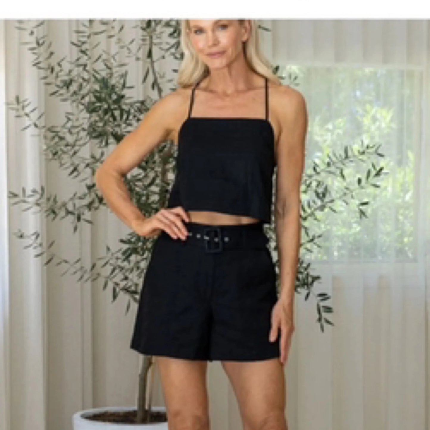 Woman wearing a black outfit with a belt in front of a decorative plant and curtain.