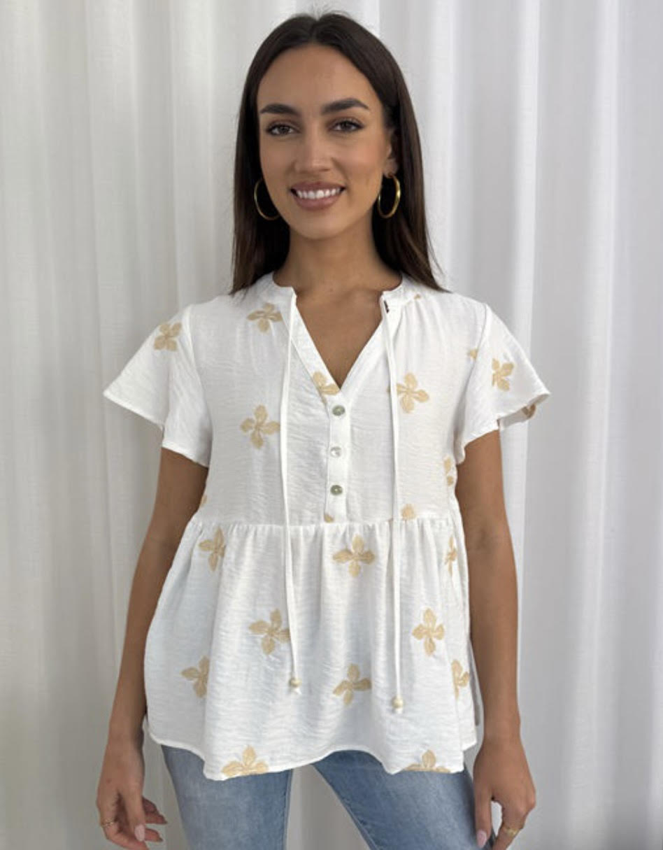 Woman wearing a white blouse with gold floral patterns against a white curtain background