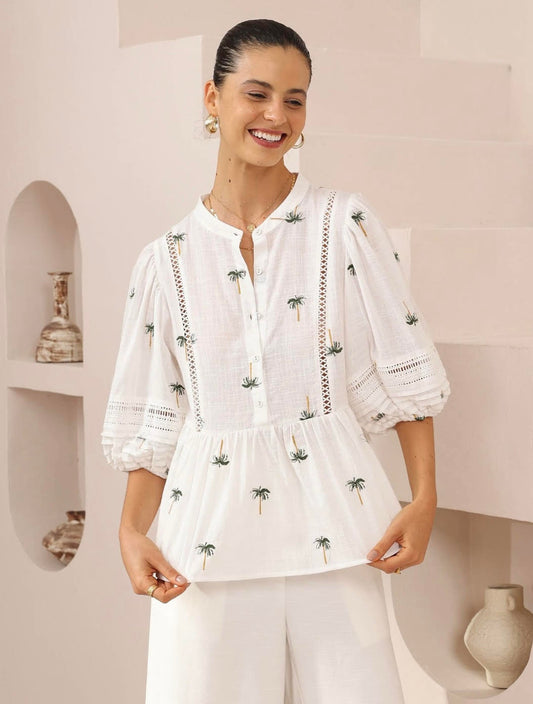 Woman wearing a white blouse with green patterns in a minimalistic room.