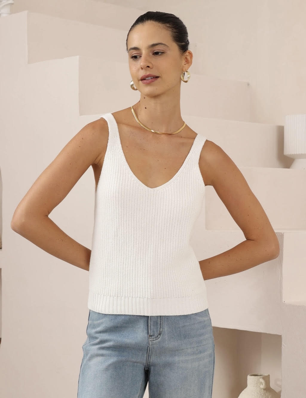 Woman wearing a white ribbed tank top and light blue jeans indoors.