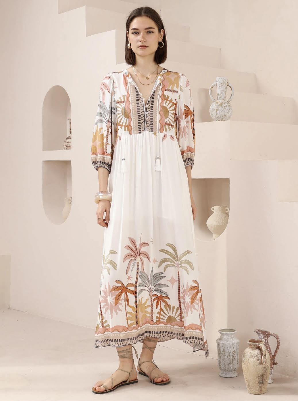 Woman wearing a long, patterned dress standing in a minimalistic room with decorative vases.