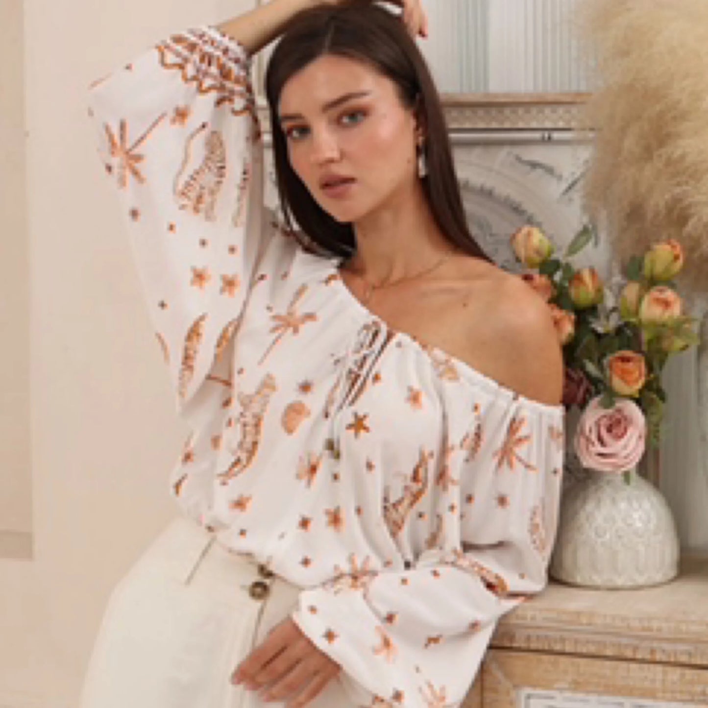 Woman wearing a white off-shoulder top with floral patterns, standing next to a vase of flowers.