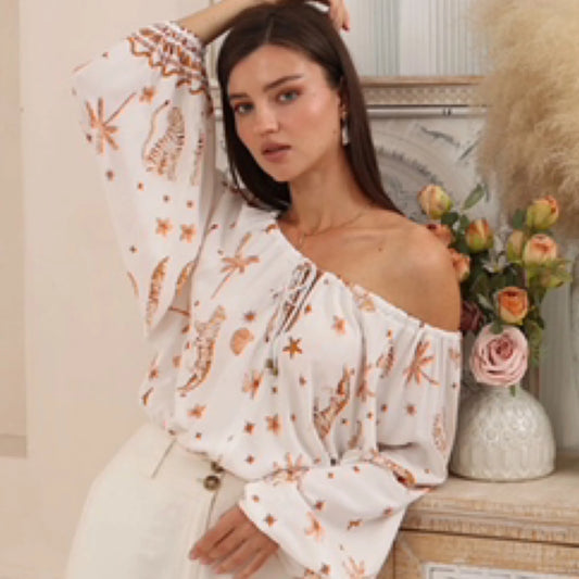 Woman wearing a white off-shoulder top with floral patterns, standing next to a vase of flowers.
